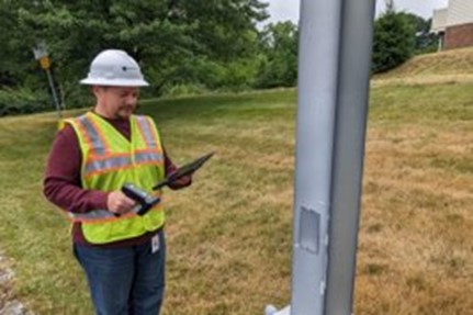 An image of a PennDOT worker in a white hard hat and yellow safety vest with a mobile device in one hand and a radio-frequency identification (RFID) reader in the other hand scanning a sign along a roadway